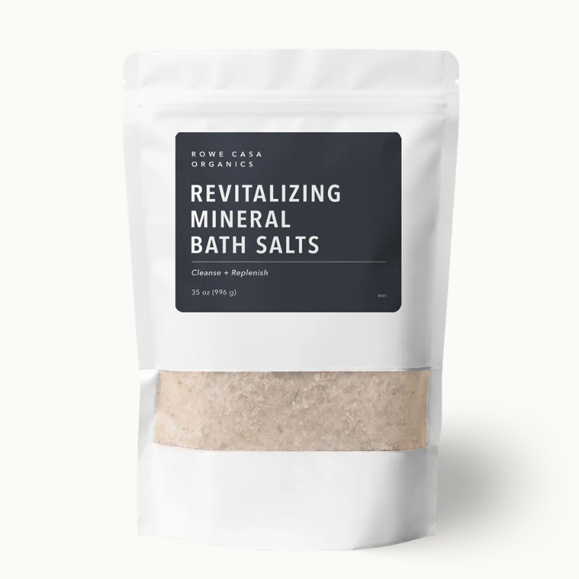 A white stand-up pouch of Rome Casa Organics Revitalizing Mineral Bath Salts, featuring a black label and a clear window showing the beige bath salts inside, set against a clean off-white background.