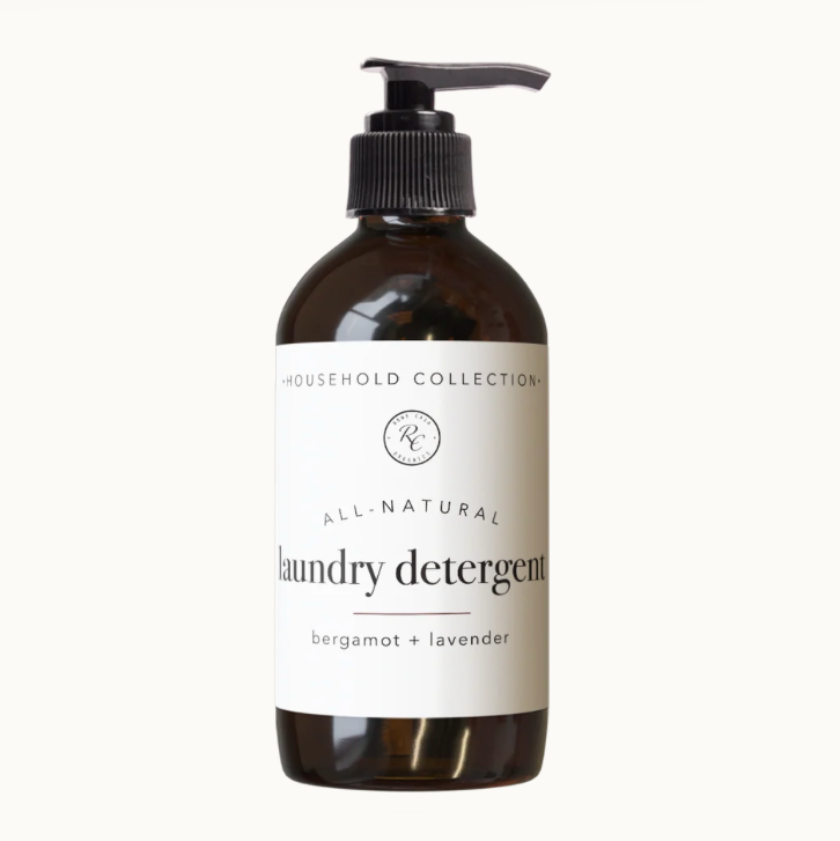 A dark amber glass bottle of laundry detergent with a black pump dispenser and a white label. The label reads 'HOUSEHOLD COLLECTION', 'ALL-NATURAL', and 'laundry detergent' with 'bergamot + lavender' underneath.