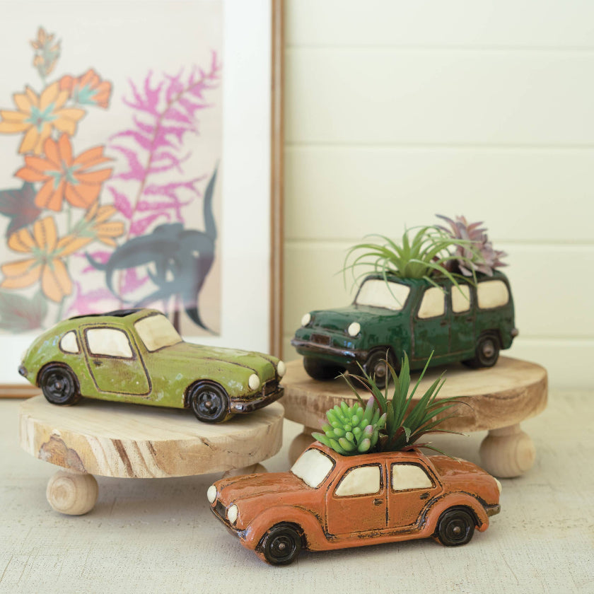 Three toy cars repurposed as planters with plants on a wooden surface, framed artwork in the background.
