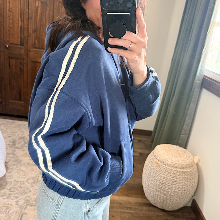 A person taking a mirror selfie wearing a navy blue pullover hoodie with white double stripes down the sleeves, paired with light-wash jeans in a home setting.