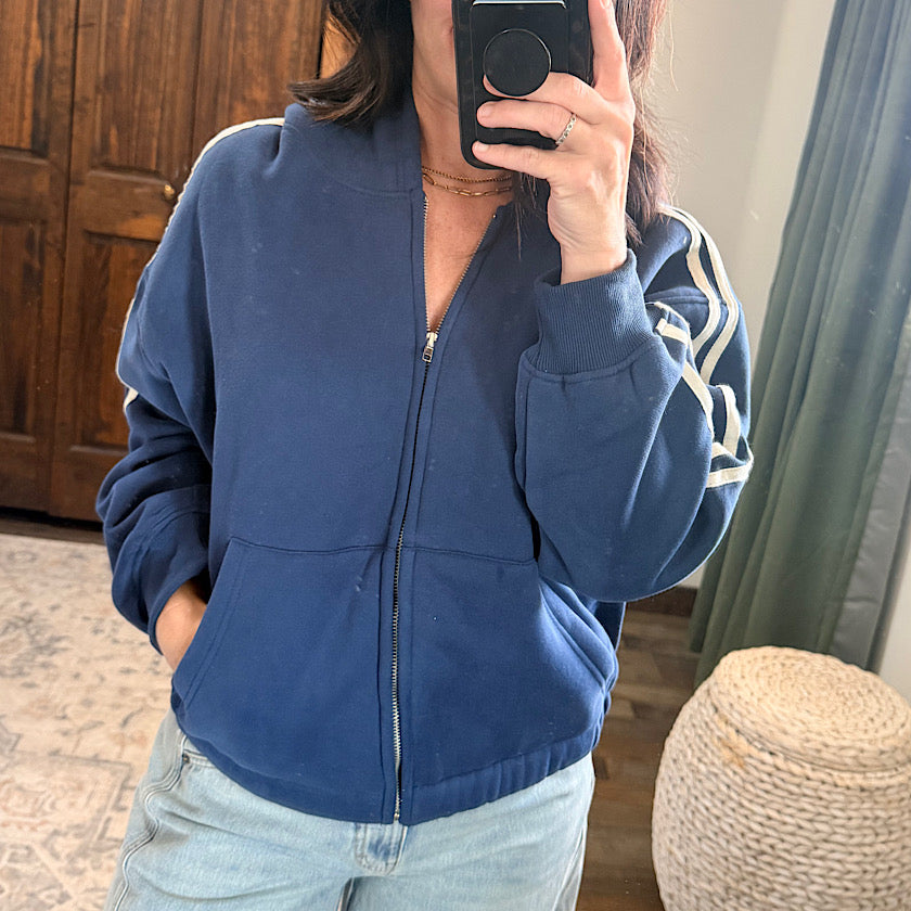 A person taking a mirror selfie wearing a navy blue zip-up hoodie with white stripes on the sleeves, paired with light-wash jeans in an indoor room setting.