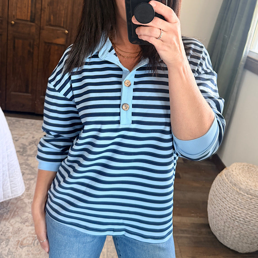 A woman takes a mirror selfie wearing a light blue and navy blue horizontally striped polo shirt with a light blue collar and cuffs, paired with blue jeans.