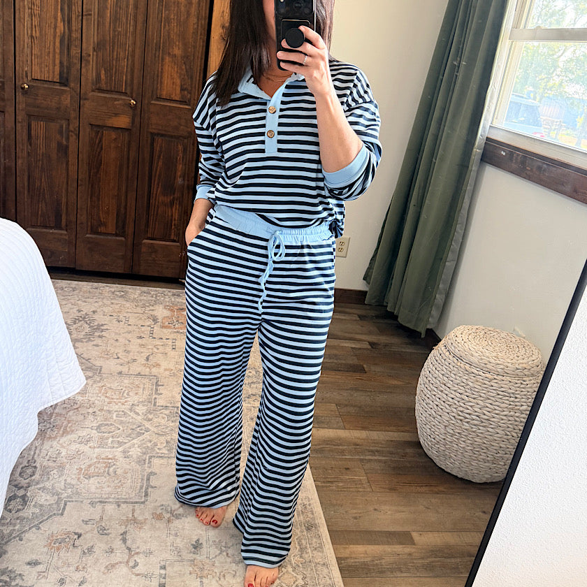 A woman takes a mirror selfie in a bedroom while wearing a matching two-piece loungewear set with black and light blue horizontal stripes. The set includes a long-sleeve henley-style top and wide-leg pants with a drawstring waist.