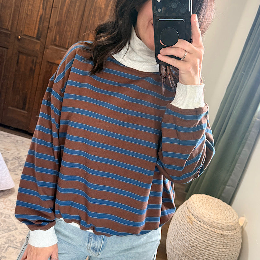 A person taking a mirror selfie wearing a brown and blue horizontally striped long-sleeve top over a white turtleneck, paired with light blue jeans in a home setting.
