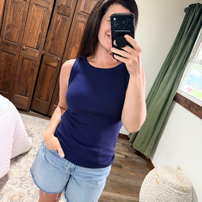 Woman taking a mirror selfie wearing a blue tank top and light blue shorts in a room with wooden flooring and green curtains.