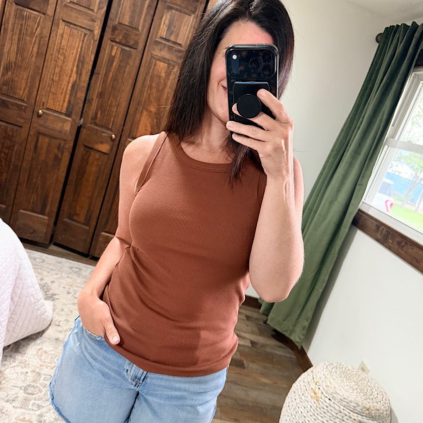 Woman taking a mirror selfie wearing a brown tank top and light blue jeans in a room with wooden cabinets and green curtains.