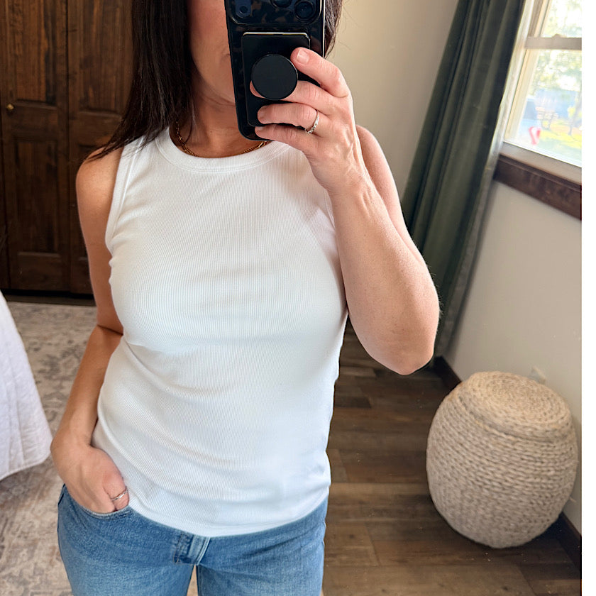 A woman takes a mirror selfie wearing a fitted white ribbed tank top and blue jeans in a home setting with wood floors, a woven pouf, and a window in the background.