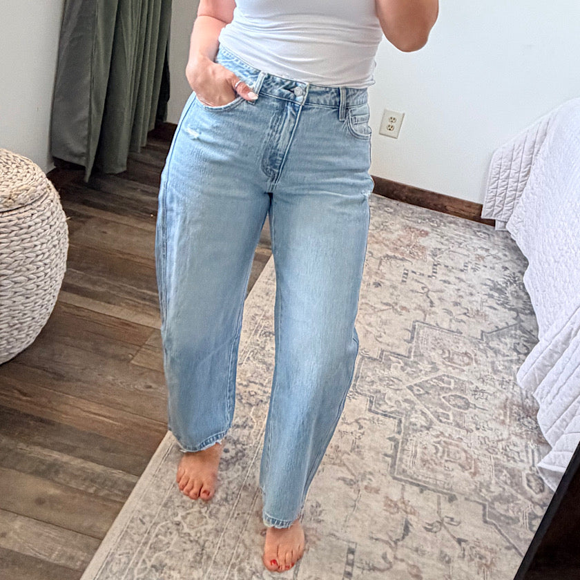 A person wearing light-wash blue straight-leg jeans and a white top, standing barefoot on a patterned rug in a bedroom setting.