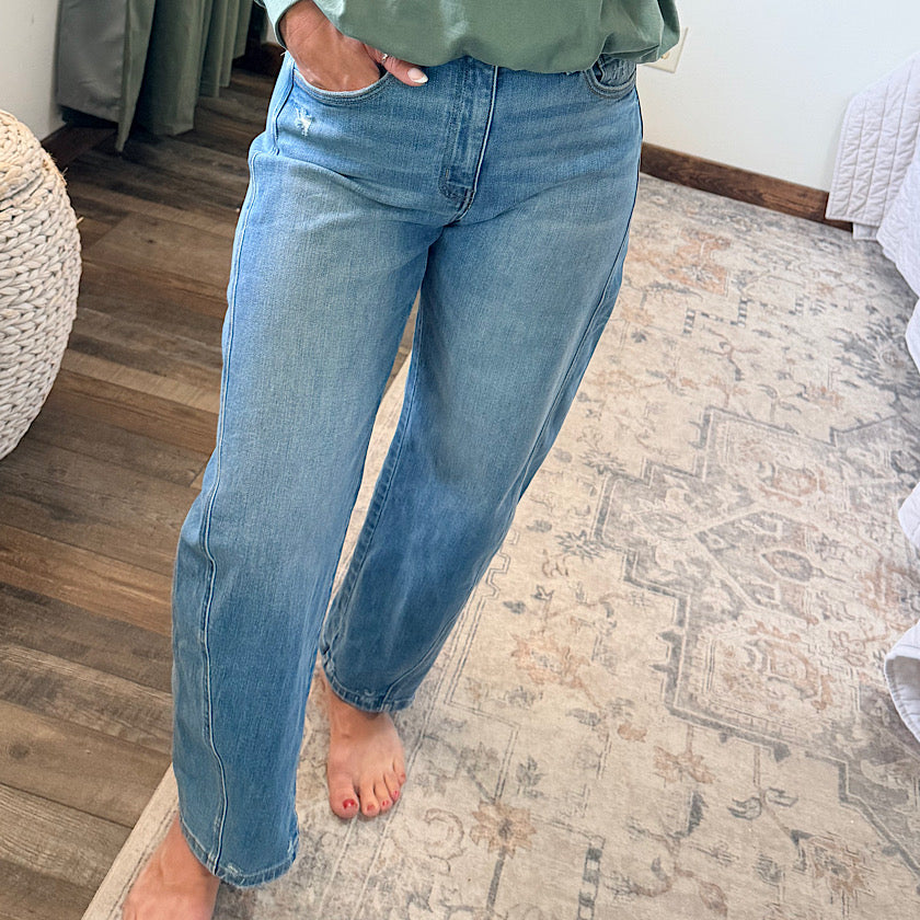 A person wearing light blue straight-leg jeans, standing barefoot on a patterned area rug in a bedroom setting.