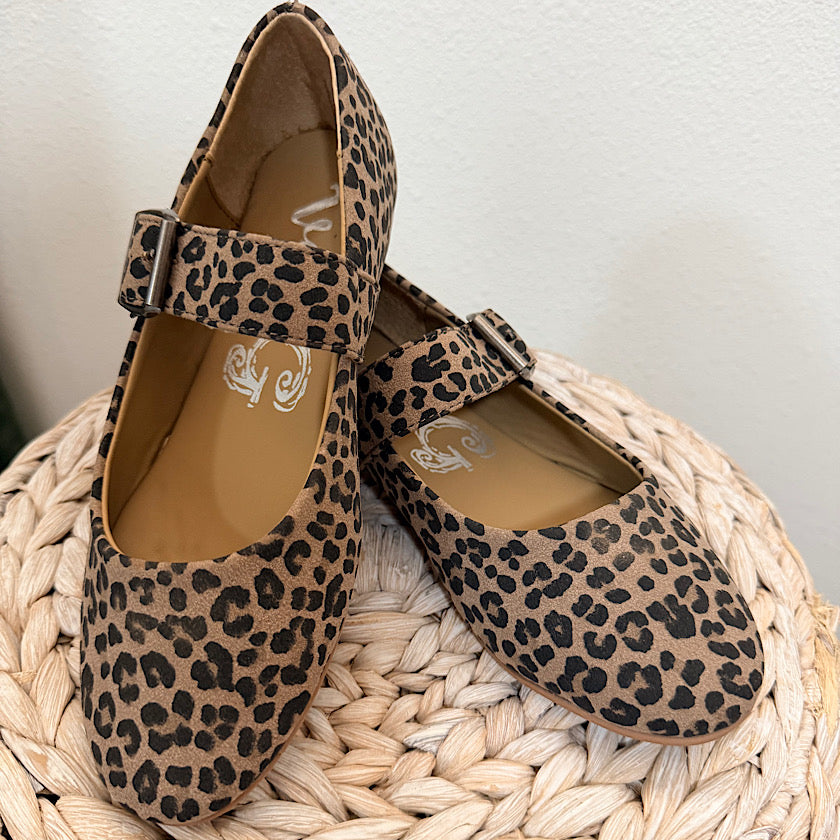 A pair of women's Mary Jane flats with a tan and black leopard print, resting on a woven straw stool against a white wall.