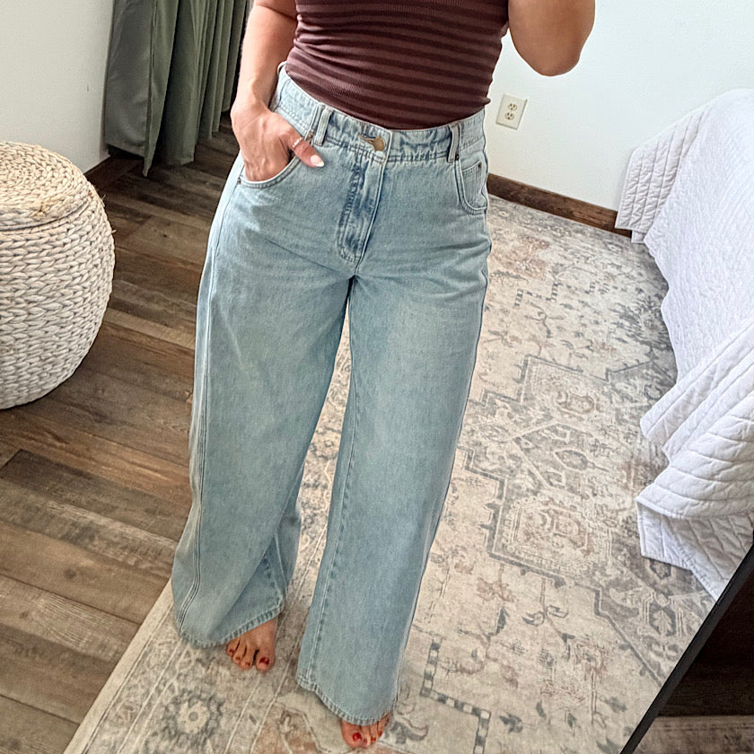 A person wearing light-wash wide-leg denim jeans and a brown striped top, standing barefoot on a patterned rug in a bedroom setting.