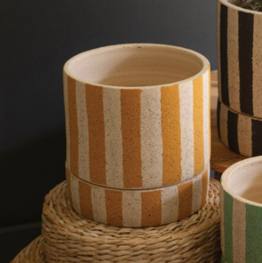 Three striped woven planters in different sizes arranged on a round woven stool beside a wooden side table, with a potted plant in the tallest planter against a dark gray wall.