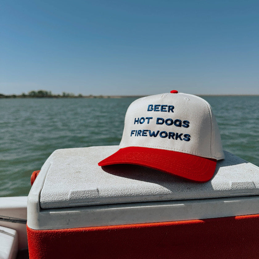 Cap with 'Beer Hot Dogs Fireworks' text on a cooler by a body of water