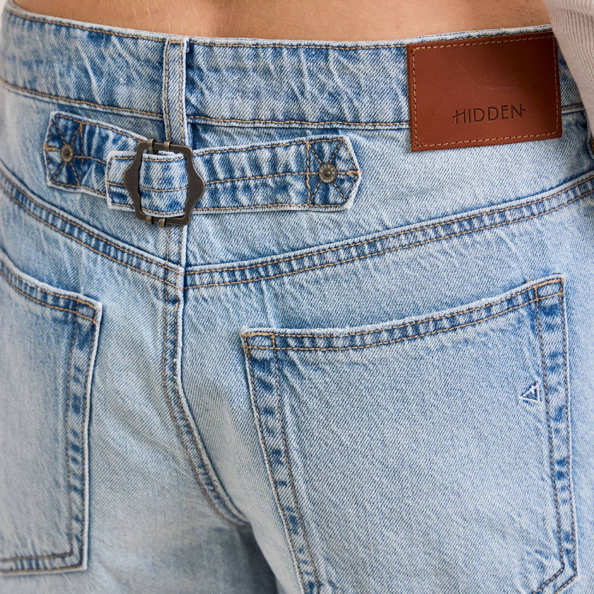 Close-up of light blue denim shorts with a brown leather patch and 'HIDDEN' branding.